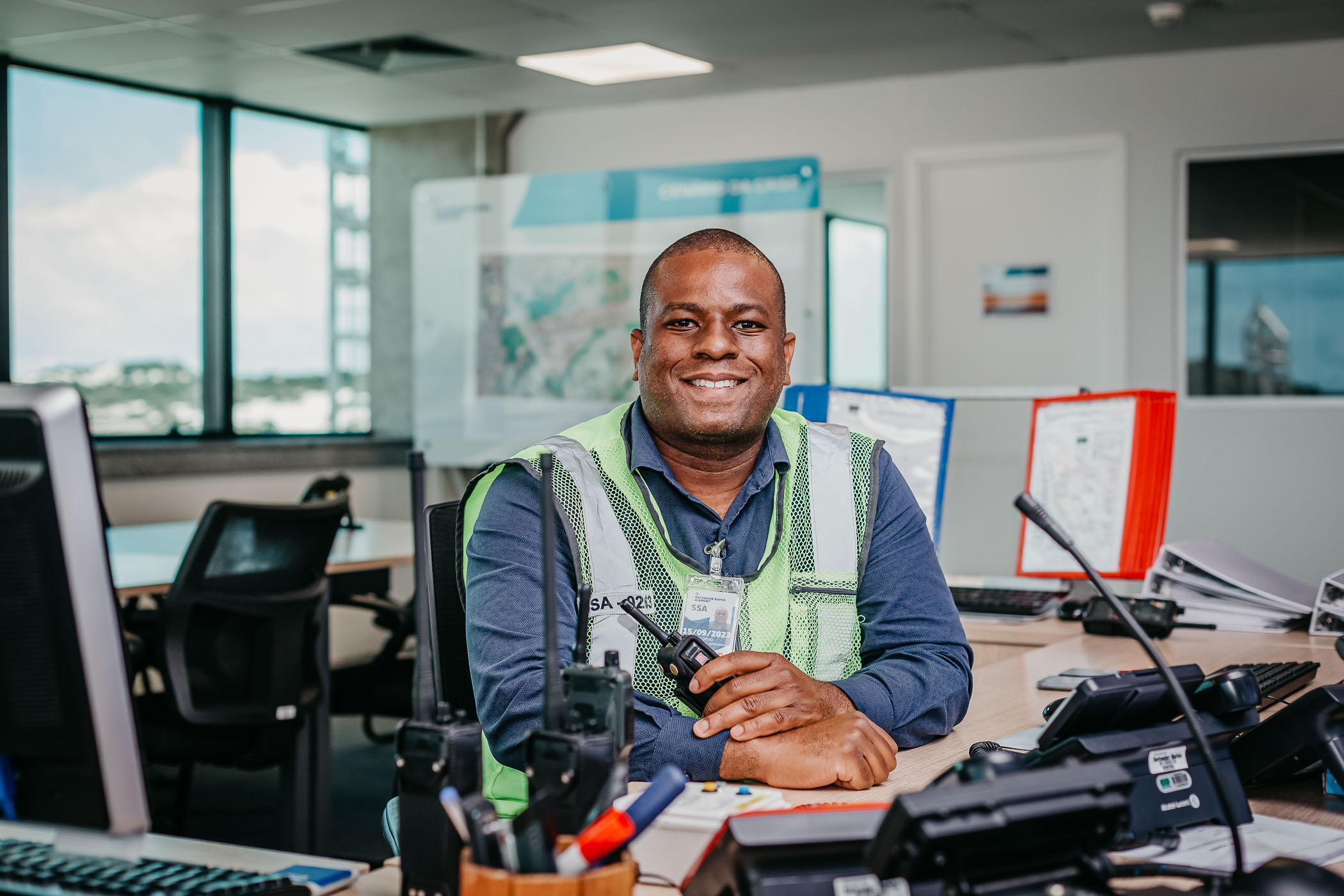 Homem negro, adulto, sentado em uma mesa de trabalho em um ambiente operacional. Ele sorri para a câmera, usando camisa azul, colete refletivo verde e crachá de identificação. Em uma das mãos, segura um rádio comunicador. Sobre a mesa há equipamentos de comunicação, papéis e um teclado. Ao fundo, aparecem cadeiras, monitores e janelas com luz natural, indicando um espaço profissional e organizado.